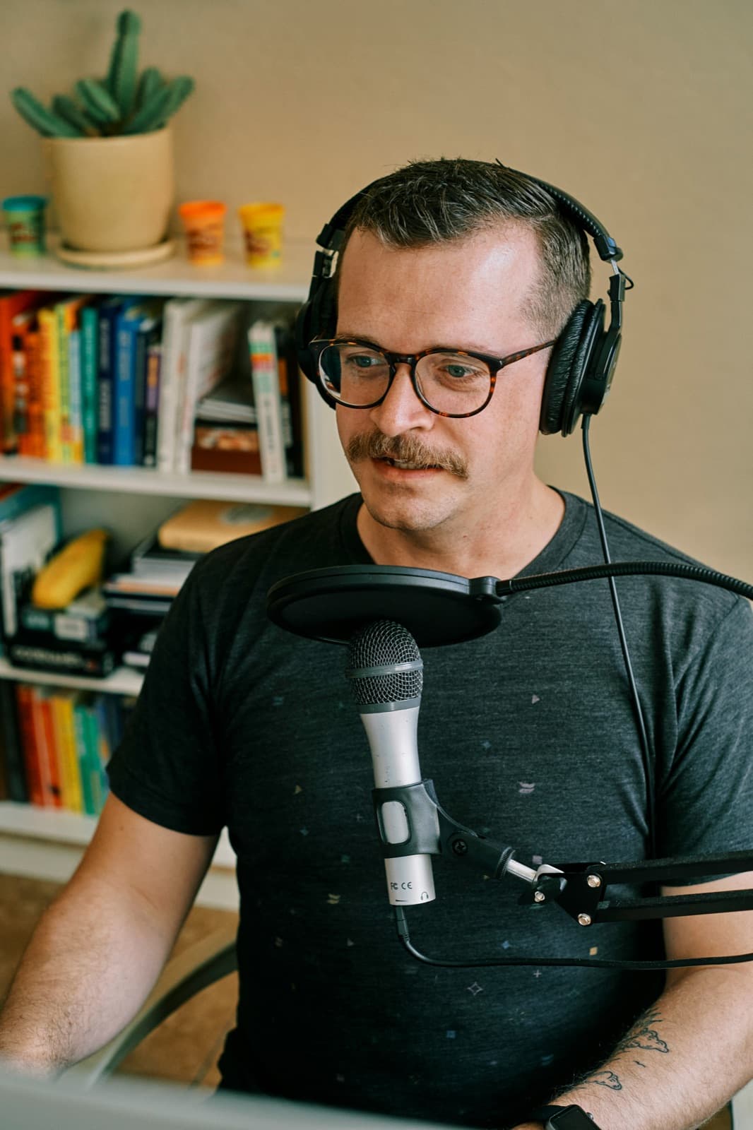 A creator wearing headphones speaking into a studio microphone at a desk, with a bookshelf of colourful books behind them.
