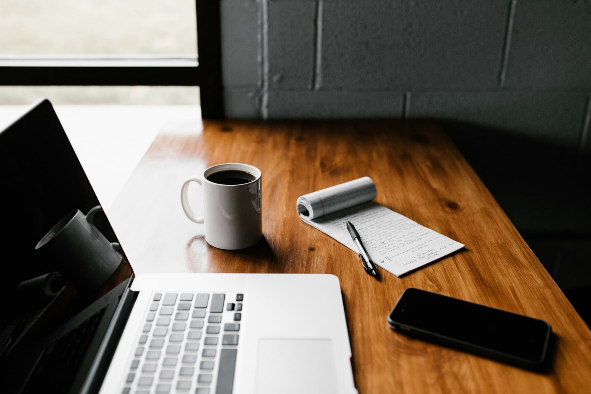 An empty wooden desk beside a window, with an open laptop, a cup of coffee, a notepad and a phone — nobody sitting at it.
